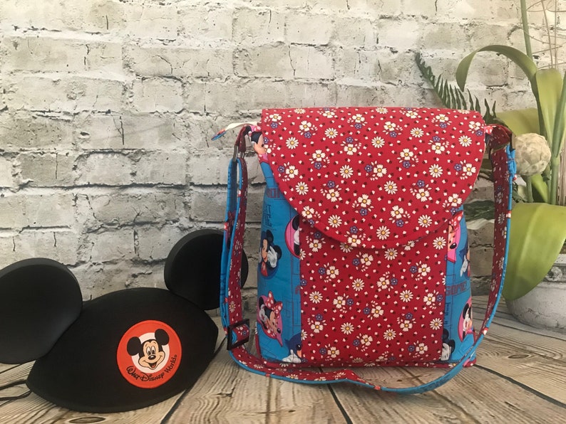 Puede incluir: Un bolso bandolera de tela azul y rojo con estampado floral y cierre de solapa. El bolso presenta un dise&ntilde;o de Mickey Mouse y Minnie Mouse. El bolso est&aacute; sobre una superficie de madera con un gorro de orejas de Mickey Mouse negro.