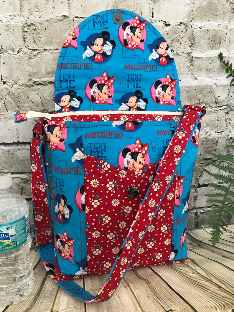 Puede incluir: Un bolso bandolera de tela azul y roja con cierre de solapa. El bolso presenta un estampado floral y un estampado repetido de personajes de Mickey Mouse y Minnie Mouse con el texto "Text Me" y "Awesome!" impreso en la tela.