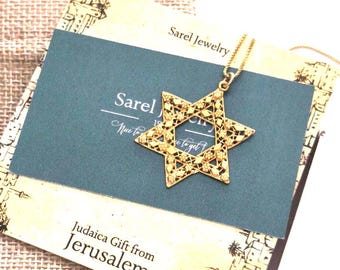 May include: A gold-colored Star of David pendant necklace. The pendant is intricately designed and rests on a teal card with the text "Sarel Jewelry" and "Judaica Gift from Jerusalem." The necklace is displayed with a burlap gift bag.