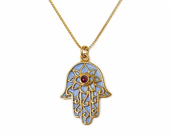 May include: A gold-tone necklace featuring a Hamsa hand pendant with light blue enamel inlay and a small, round, dark red stone set in the center of the eye motif. The pendant hangs from a delicate gold-tone box chain with a polished finish.