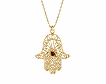 May include: A gold-toned necklace featuring a Hamsa hand pendant with intricate filigree metalwork and a single small, round, dark red gemstone set in the center of the palm. The pendant hangs from a delicate, matching gold-toned box chain.