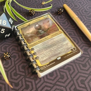 Puede incluir: Una carta de Magic: The Gathering amarilla y dorada con Aminatou, la Forjadora del Destino, una planeswalker legendaria. La carta tiene una imagen de Aminatou, una mujer con un tocado blanco, sosteniendo un arco y una flecha. El texto de la carta describe las habilidades de Aminatou.