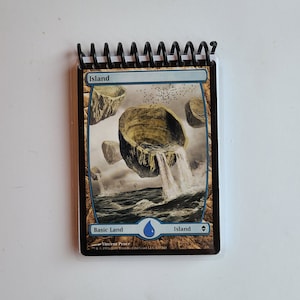 Puede incluir: Un pequeño bloc de notas con espiral que presenta una ilustración de una carta de Magic: The Gathering. La carta muestra un paisaje de fantasía con islas flotantes, cascadas y la palabra "Island". Es una carta de tierra básica.