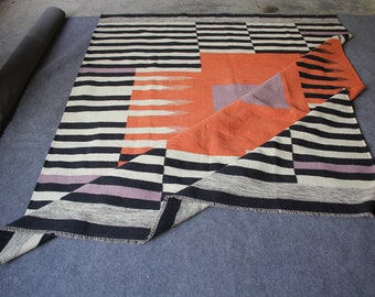 May include: A geometric patterned rug with a folded corner. The rug features black and white stripes, an orange central section, and accents of purple. The rug is made of woven fabric and is displayed on a gray surface.