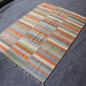 May include: A rectangular rug with a striped pattern in shades of orange, blue, green, and gray. The rug has a fringe border.
