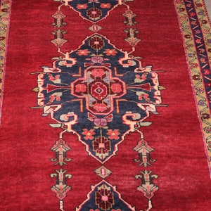 K&ouml;nnte beinhalten: Ein roter Teppich mit einem geometrischen Muster in Marineblau und Creme. Der Teppich hat einen Rand mit einem floralen Muster in Rot-, Blau- und Cremet&ouml;nen.