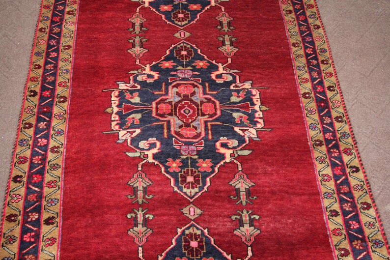 K&ouml;nnte beinhalten: Ein roter Teppich mit einem geometrischen Muster in Marineblau und Creme. Der Teppich hat einen Rand mit einem floralen Muster in Rot-, Blau- und Cremet&ouml;nen.
