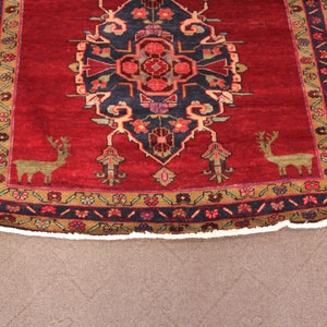 K&ouml;nnte beinhalten: Ein roter Teppich mit traditionellem persischem Design, das ein zentrales Medaillon mit stilisierten floralen Motiven und zwei Hirschen in den unteren Ecken zeigt. Der Teppich hat einen dunkelblauen Rand mit einem geometrischen Muster.