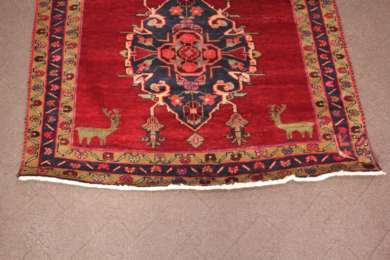 K&ouml;nnte beinhalten: Ein roter Teppich mit traditionellem persischem Design, das ein zentrales Medaillon mit stilisierten floralen Motiven und zwei Hirschen in den unteren Ecken zeigt. Der Teppich hat einen dunkelblauen Rand mit einem geometrischen Muster.