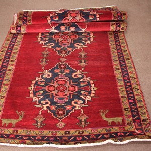 K&ouml;nnte beinhalten: Ein roter, blauer und goldfarbener Teppich mit Fransen. Der Teppich hat ein traditionelles Design mit floralen Motiven und zwei stilisierten Hirschen.
