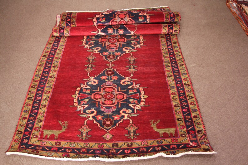 K&ouml;nnte beinhalten: Ein roter, blauer und goldfarbener Teppich mit Fransen. Der Teppich hat ein traditionelles Design mit floralen Motiven und zwei stilisierten Hirschen.