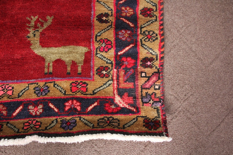 K&ouml;nnte beinhalten: Ein roter Teppich mit einem braunen und blauen Rand. Der Teppich hat ein stilisiertes Hirsch-Design in der Mitte. Der Teppich hat einen Fransenrand.