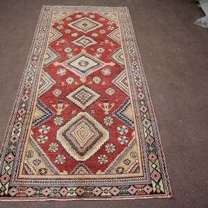 May include: A red, beige, and blue patterned rug with a geometric design. The rug has a fringe border.