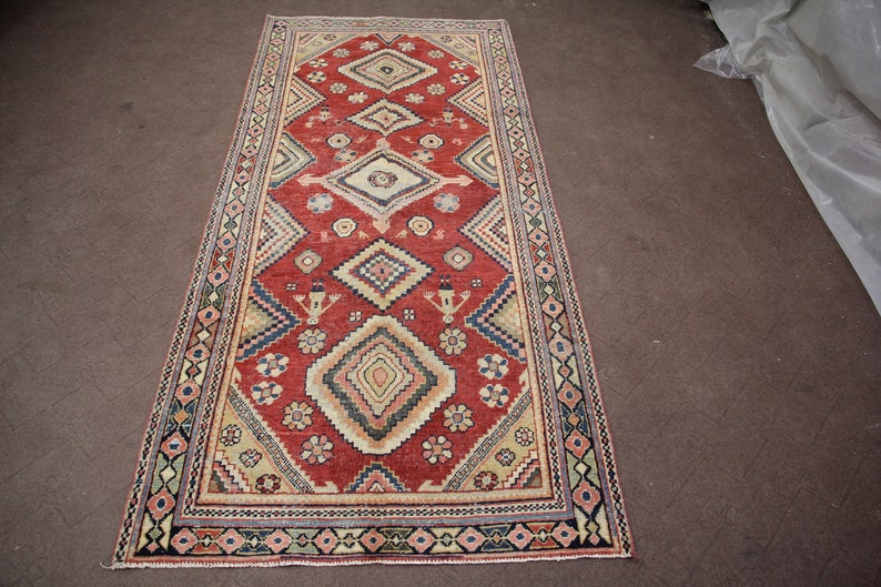 May include: A red, beige, and blue patterned rug with a geometric design. The rug has a fringe border.