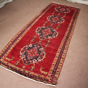 K&ouml;nnte beinhalten: Ein roter L&auml;ufer mit traditionellem persischem Design. Der Teppich hat eine dunkelblaue Mitte mit einem Blumenmuster und einen roten Rand mit einem geometrischen Muster.