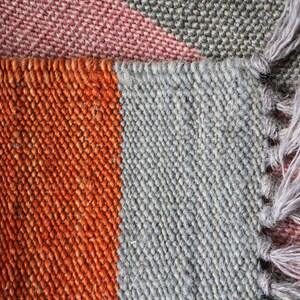 May include: Close-up of a woven rug with stripes of orange, gray, and pink. The rug has a fringe on one side.