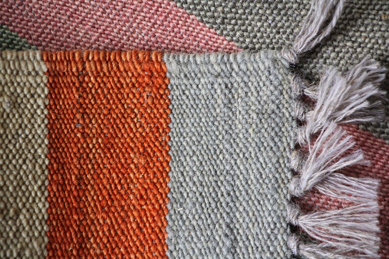 May include: Close-up of a woven rug with stripes of orange, gray, and pink. The rug has a fringe on one side.