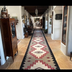 May include: A long hallway with a Southwestern-style rug featuring a geometric pattern in red, black, and cream. The walls are white, and the floor is a light brown stone. Antique furniture and artwork adorn the walls.