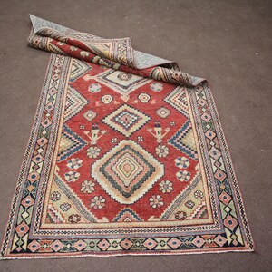 May include: A red, beige, and blue patterned rug with a geometric design. The rug has a fringe border.