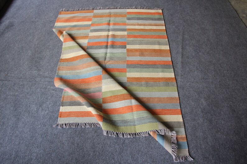 May include: A multi-colored striped rug with a fringe border. The rug is folded in half and the colors are orange, blue, green, and beige.