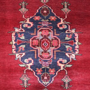 K&ouml;nnte beinhalten: Ein roter Teppich mit einem blauen und wei&szlig;en floralen Muster in der Mitte. Der Teppich hat einen Rand mit einem geometrischen Muster in Braun-, Blau- und Beiget&ouml;nen.