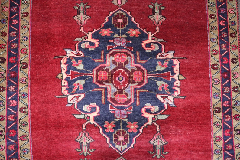 K&ouml;nnte beinhalten: Ein roter Teppich mit einem blauen und wei&szlig;en floralen Muster in der Mitte. Der Teppich hat einen Rand mit einem geometrischen Muster in Braun-, Blau- und Beiget&ouml;nen.