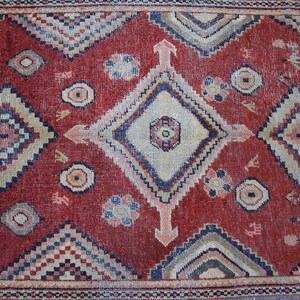May include: A red rug with a geometric pattern in shades of blue, beige, and white. The rug has a central diamond shape with smaller diamond shapes surrounding it.