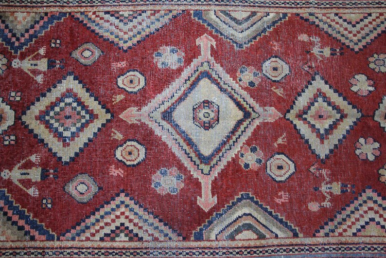 May include: A red rug with a geometric pattern in shades of blue, beige, and white. The rug has a central diamond shape with smaller diamond shapes surrounding it.