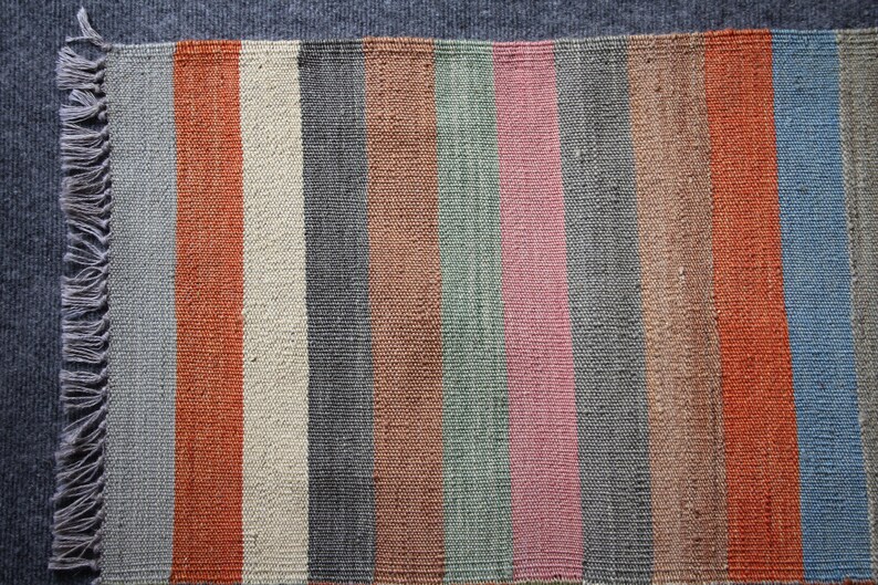 May include: A striped woven rug with a fringe on one side. The rug has alternating stripes of gray, orange, beige, brown, green, pink, and blue.