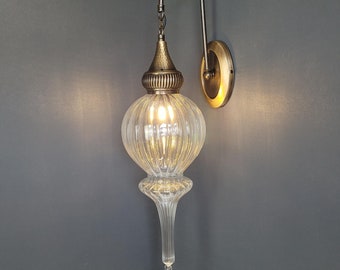 May include: A clear glass wall sconce with a brass finish and a decorative metal finial. The sconce has a round, ribbed glass shade and a curved arm.