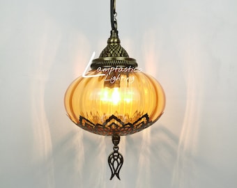 May include: A amber glass pendant light with a decorative metal cage and a chain hanging from the ceiling. The light is on and the bulb is glowing. The text "Lampfastics Lighting" is visible on the glass.