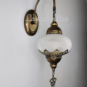 May include: A white frosted glass wall sconce with a gold metal frame and ornate details. The sconce has a curved arm and a chain hanging from the top.