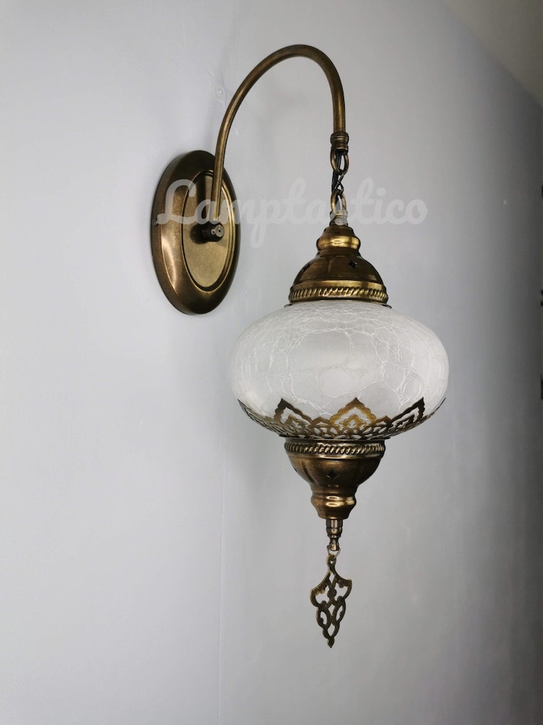 May include: A white frosted glass wall sconce with a gold metal frame and ornate details. The sconce has a curved arm and a chain hanging from the top.
