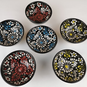 May include: Six small black ceramic bowls with white, blue and yellow floral designs. The bowls are all the same size and shape, and they are arranged in a circle on a white background.