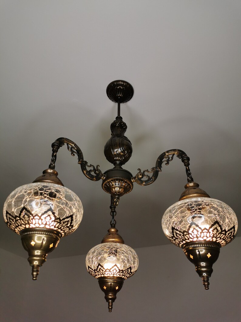 May include: A three-light chandelier with a black metal frame and clear glass shades with intricate metalwork. The shades have a crackled glass effect and are illuminated by light bulbs.