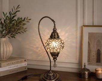 May include: A table lamp with a curved bronze-toned metal stand and a spherical glass shade featuring a white geometric mosaic pattern. The lamp emits a warm glow, casting intricate star-shaped shadows on the wall behind it.