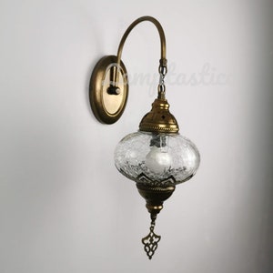 May include: A gold-toned wall sconce with a clear, textured glass shade. The sconce has a curved arm and a decorative chain hanging from the bottom of the shade.