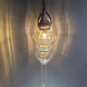 May include: A clear glass pendant light fixture with a decorative metal top and hanging chain. The light has a bulb inside, illuminating the ribbed glass design. The fixture has a long, tapered bottom and a small decorative charm.
