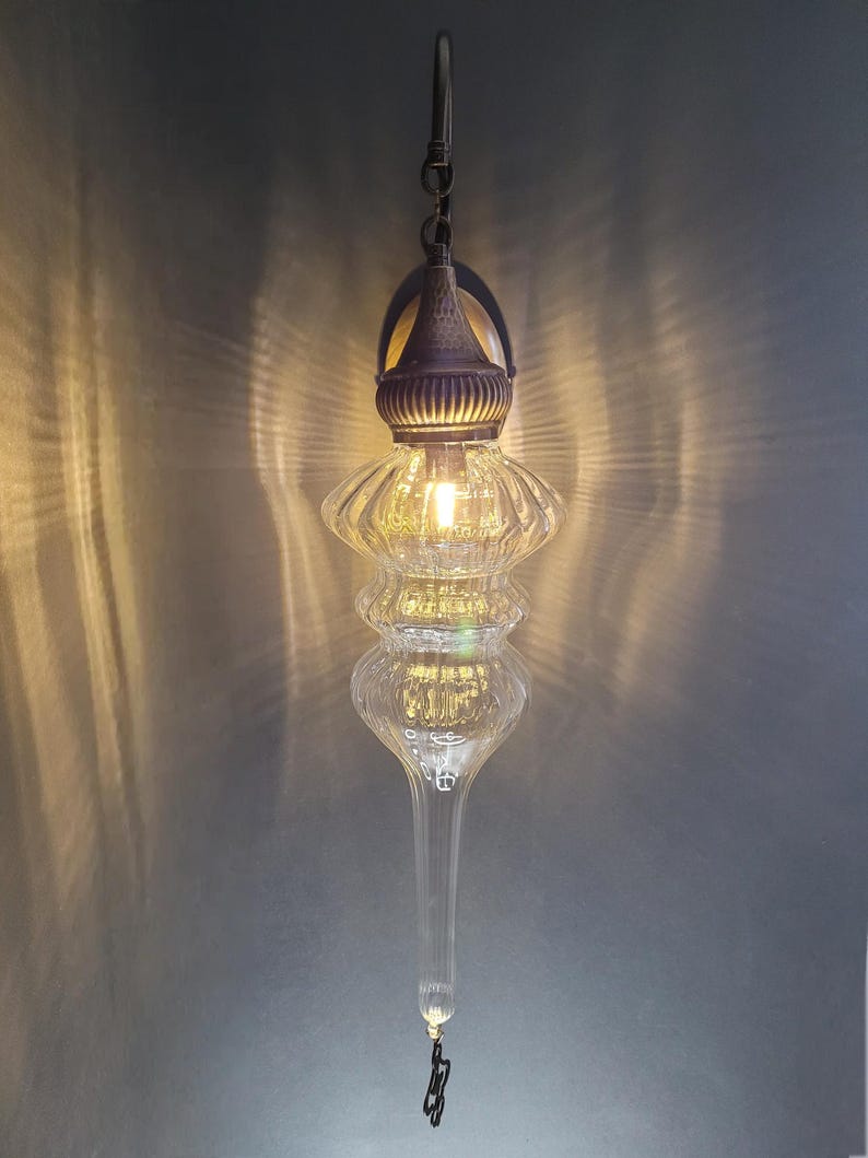 May include: A clear glass pendant light fixture with a decorative metal top and hanging chain. The light has a bulb inside, illuminating the ribbed glass design. The fixture has a long, tapered bottom and a small decorative charm.