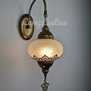 May include: A white frosted glass Turkish style wall sconce with a brass finish and intricate metalwork. The sconce has a curved arm and a decorative hanging ornament.
