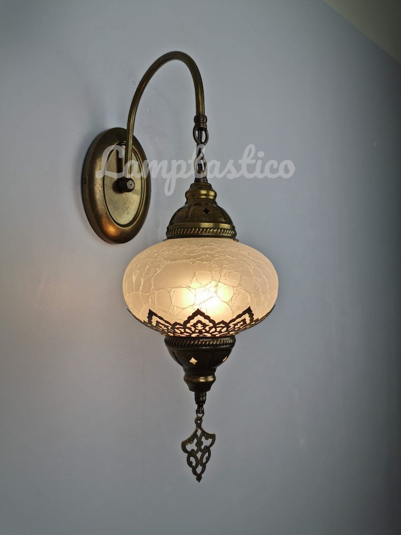 May include: A white frosted glass Turkish style wall sconce with a brass finish and intricate metalwork. The sconce has a curved arm and a decorative hanging ornament.