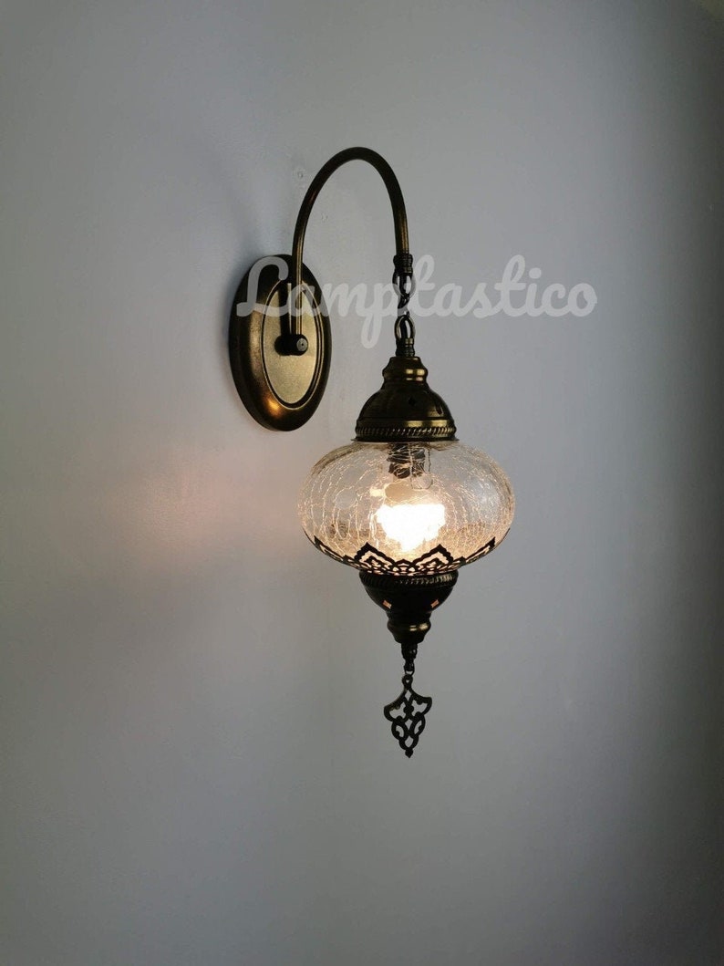 May include: A wall-mounted light fixture with a curved arm and a clear glass globe shade. The shade has a textured surface and a decorative metal finial. The fixture is finished in a brass tone.