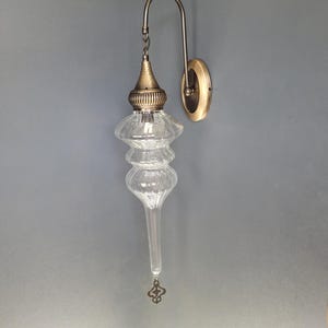 May include: An antique-style wall sconce with a clear, teardrop-shaped glass shade and a decorative brass-colored metal frame. The fixture has a curved arm and a round wall plate. The glass shade has a textured surface and a decorative finial.