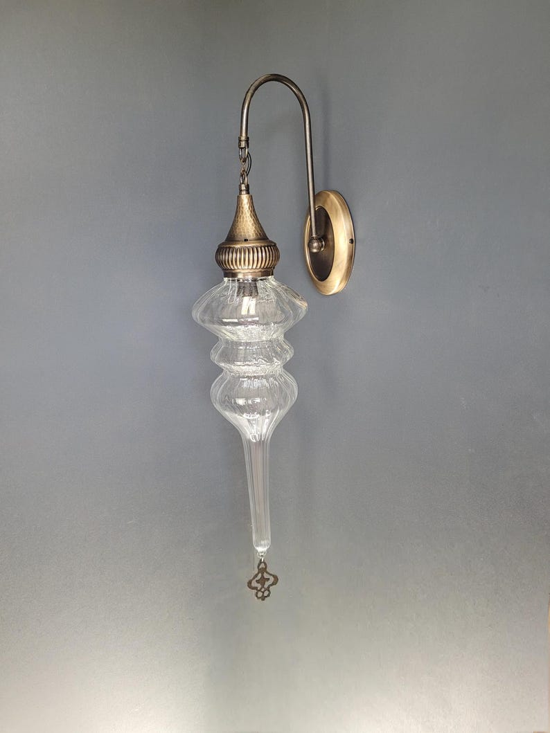 May include: An antique-style wall sconce with a clear, teardrop-shaped glass shade and a decorative brass-colored metal frame. The fixture has a curved arm and a round wall plate. The glass shade has a textured surface and a decorative finial.