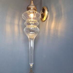 May include: An ornate wall sconce with a clear, elongated glass shade and a decorative brass-colored metal fixture. The light fixture has a bulb inside, and is mounted on a wall with a brass-colored circular base.
