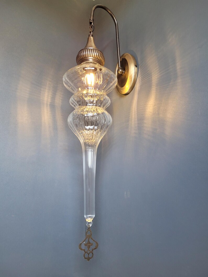 May include: An ornate wall sconce with a clear, elongated glass shade and a decorative brass-colored metal fixture. The light fixture has a bulb inside, and is mounted on a wall with a brass-colored circular base.