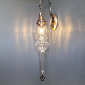 May include: A clear glass wall sconce with a decorative metal fixture. The sconce has a long, teardrop-shaped glass body with a textured design. The metal components are a brass color, and the light is on.