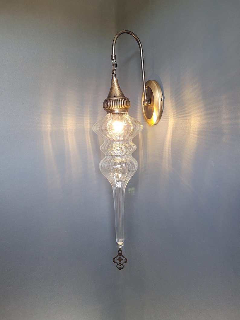 May include: A clear glass wall sconce with a decorative metal fixture. The sconce has a long, teardrop-shaped glass body with a textured design. The metal components are a brass color, and the light is on.