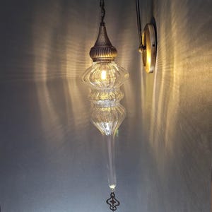 May include: A clear glass wall sconce with a decorative metal fixture. The sconce has a bulbous, ribbed glass shade and a long, tapered bottom. The metal arm and wall plate have a brass finish. The light is on, casting a warm glow.