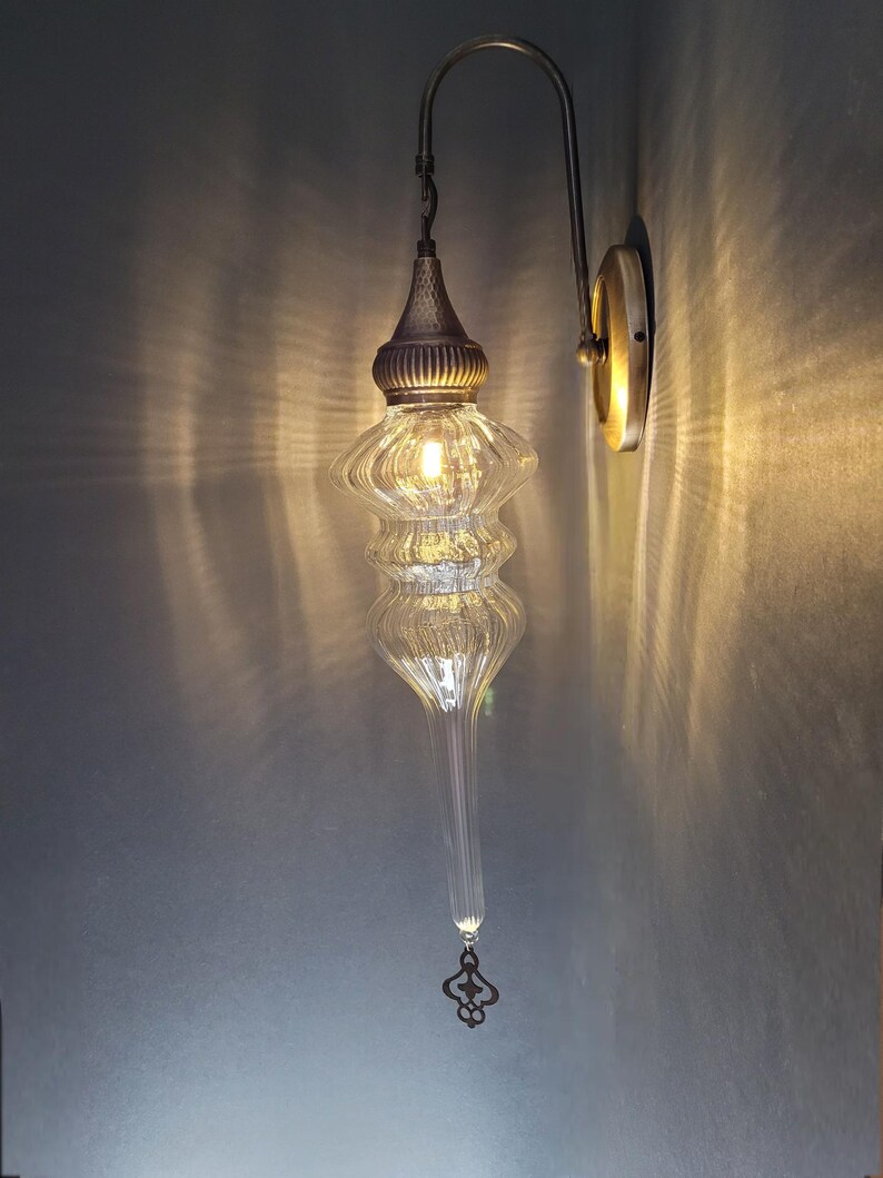 May include: A clear glass wall sconce with a decorative metal fixture. The sconce has a bulbous, ribbed glass shade and a long, tapered bottom. The metal arm and wall plate have a brass finish. The light is on, casting a warm glow.